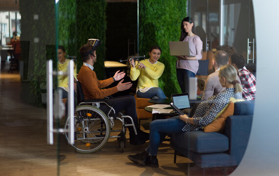 Disabled Businessman In A Wheelchair At Work In Modern Open Space Coworking Office With Team Using Virtual Reality Googles Drone Assistance Simulation