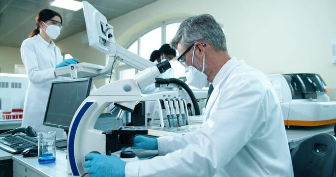 Side view of senior Caucasian male medical researcher working in lab on microscope equipment and typing on laptop at workplace, Healthcare worker checking analysis in laboratory, microbiology concept