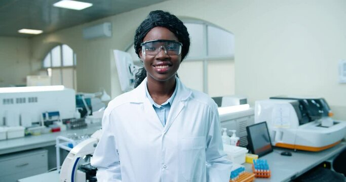 Close up portrait of happy positive young African American beautiful female medical scientist in goggles and white coat standing in clinic lab and smiling looking at camera Modern laboratory, medicine