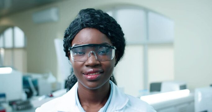Close up of young African American cheerful beautiful woman medical scientist in goggles in clinic lab, smiling looking at camera at work. Modern laboratory, medicine, healthcare worker