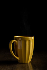 Yellow mug with hot steam rising from it while sitting in front of a black background on a dark wooden surface.