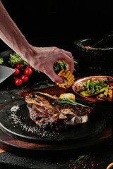 T-Bone steak serving with grilled vegetables on a black round plate on a background of greens,...