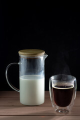 Black coffee in glass cup with double walls. Milk decanter. Wooden table and black table.
