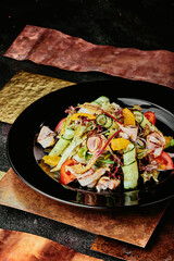 salad with ham, orange slices, tomato, cucumber and lettuce leaves in a black plate on a copper background. 