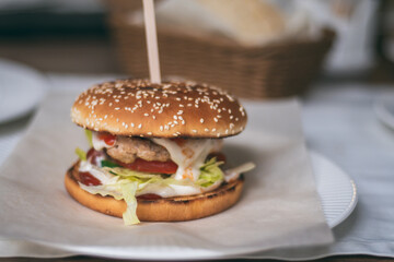 Partially blurred burger held together with wooden skewer on plate. Sesame bun, cutlet, cucumber, tomato, lettuce, sauce