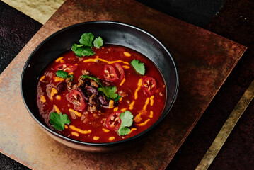 spicy tomato soup with beans, chili pepper, cheese and parsley in a black plate on a copper background