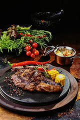 T-Bone steak serving with grilled vegetables on a black round plate on a background of greens,...