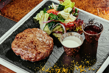 grilled meat patty with salad and two sauces on a black plate on a copper background.