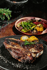 T-Bone steak serving with grilled vegetables on a black round plate on a background of greens,...