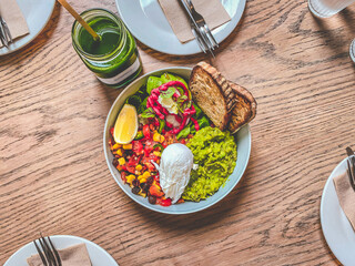 a healthy bowl of mixed vegetable salad topped with poached egg and toasts, and a jar of fresh vegetable juice on a wooden table surrounded by clean plates and cutlery. Food photography.