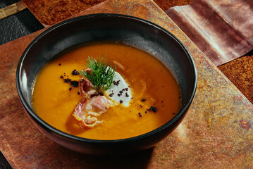 Pumpkin cream soup with bacon in a black plate on a metal background.