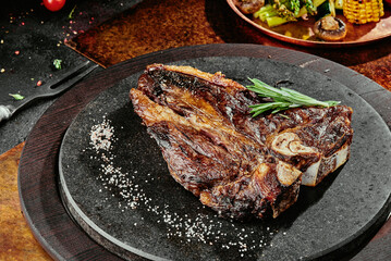T-Bone steak serving with grilled vegetables on a black round plate on a background of greens, spices and vegetables, on a copper sheet.