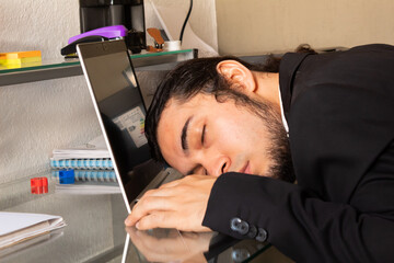 Hispanic young man in a black suit sleeping at his desk very tired from doing home office