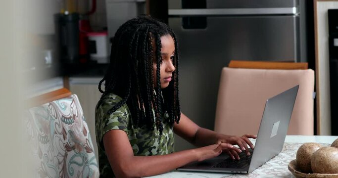 African child typing on laptop at home kitchen. Candid Black girl using computer