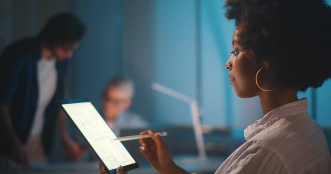 Professional afro-american architect studying blueprint on digital tablet in office working late