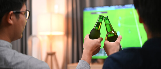Two man clinking their glasses of beer while watching football match.