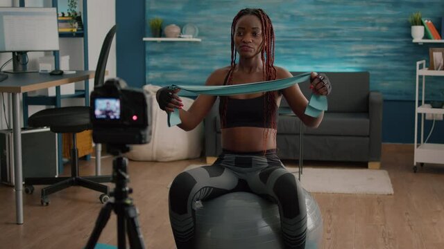 Athletic slim woman with black skin recording online yoga class using videocamera during fitness workout in living room. Fit athletic doing body exercices using aerobic elastic sitting on swiss ball