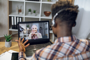 African man in headset having video chat on laptop with smiling muslim woman in hijab. Young couple communicating on distance during pandemic time.