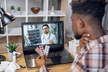Fototapeta na wymiar Young afro american guy with various pills in hands having video consultation with indian doctor while suffering from sore throat. Man in lab coat explaining to patient results of CT x-ray scan.