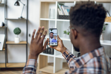 Back view of african man smiling and waving during video call with brother and nephew. Young guy using modern smartphone for online conversation with family.