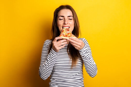 young woman with closed eyes eating a slice of hot fresh pizza on a yellow background