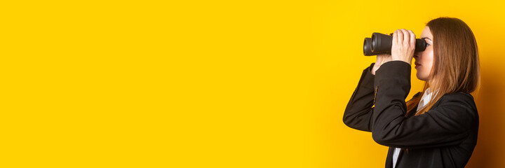 young business woman looking through binoculars on a yellow background. Banner - obrazy, fototapety, plakaty