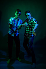 Two young men in plaid shirts dance in the studio with multicolored light