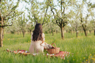 Woman is sitting on the green grass at a picnic in the orchard with basket with white flowers near.
