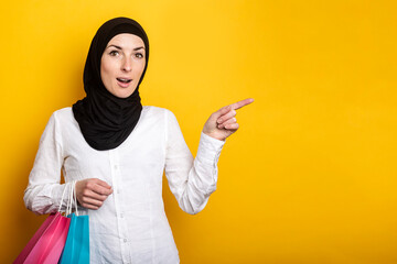 Surprised Young Muslim woman in hijab holds shopping bags and points finger to the side against yellow background. Banner