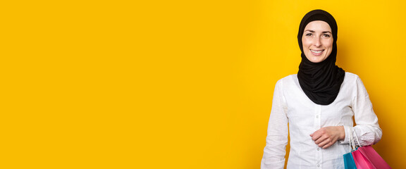 Young muslim woman in hijab smiles and holds shopping bags on yellow background. Banner