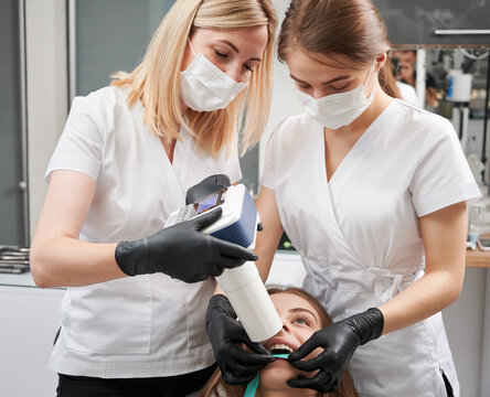 Female Dentist And Her Assistant Making X Ray Image With Portable X-ray Machine. People Wearing Clinic Uniform, Face Masks And Black Gloves. Concept Of Digital Dentistry And Intraoral Radiography.