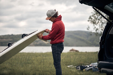 Windsurfer and camper packing and unpacking from a car's roof rack in nature.