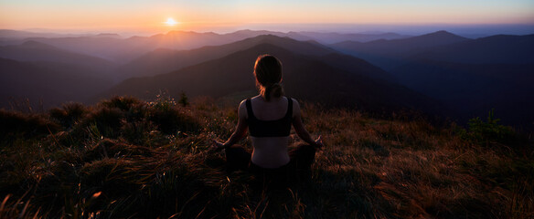 Back view of young woman in black sportswear doing meditation in the mountains at sunset. Concept of harmony with beautiful nature.