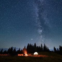 Male traveler looking at campfire while sitting near camp tent under beautiful night sky with stars...