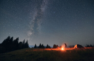 Man hiker standing on grassy hill near campfire and pointing finger at beautiful night starry sky...