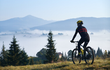 Silhouette of man cyclist in cycling suit riding bike on grassy hill. Male bicyclist enjoying the view of majestic mountains during bicycle ride. Concept of sport, bicycling and nature.