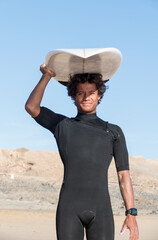 Young happy malagasy african surfer portrait holding his surfboard and smiling. Wearing black...