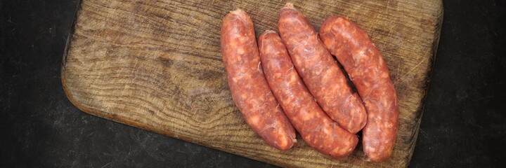 Raw Sausages In Natural Casing.  Stuffed Sausages on Old Wooden Cutting Board and Fork, Overhead View. Uncooked Sausages For Grilling or Frying on Wood Board And Black Table in Background, Top View.