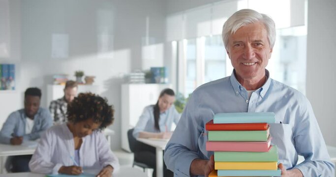 Portrait of aged male teacher holding stack of books and smiling at camera in class