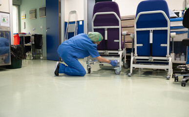 Woman from the cleaning service of a hospital crouching cleaning the wheels of a patient chair in...