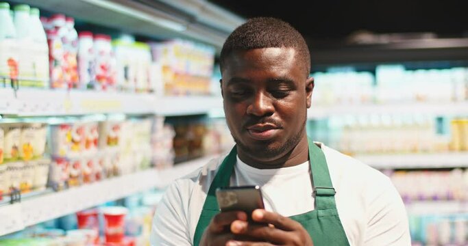Young Afro-American guy in apron using smartphone and looking at dairy prodcuts shelves. Handsome salesman scrolling feed, texting and smiling in supermarket. Social media, commerce concept.