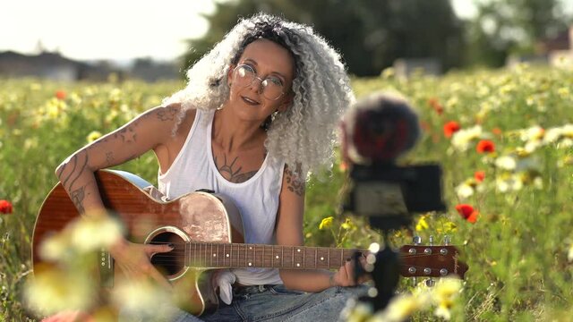 A Spanish woman with curly hair glasses and tattoos playing guitar and singing in the meadow