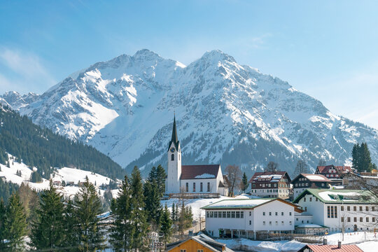Winterliche Landschaft im Kleinwalsertal mit der Pfarrkirche von Hirschegg und traditionellen H&auml;usern, umgeben von schneebedeckten Bergen der Alpen.