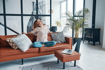 Beautiful young woman stretching and smiling while sitting on the sofa at home