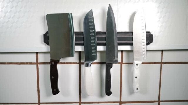 Knives set on magnet on wall in restaurant kitchen. kitchen implements hanging on magnetic holder. Chef in black gloves puts one knife. Tools for cooking. Variety of sharp knives ready for use. 4 k