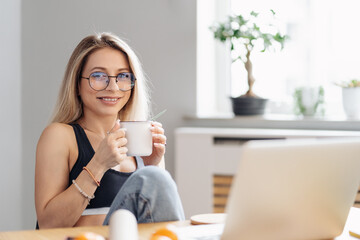 CAucasian freelancer woman work from home. Female hold tea chatting or watching movie use laptop. Modern girl relaxing at kitchen surfing internet