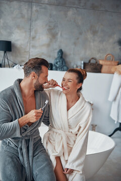 Happy young couple in bathrobes cleaning teeth while doing morning routine