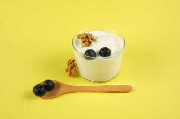 Glass cup with creamy yogurt and blueberries and walnuts with a yellow background and a wooden spoon horizontally