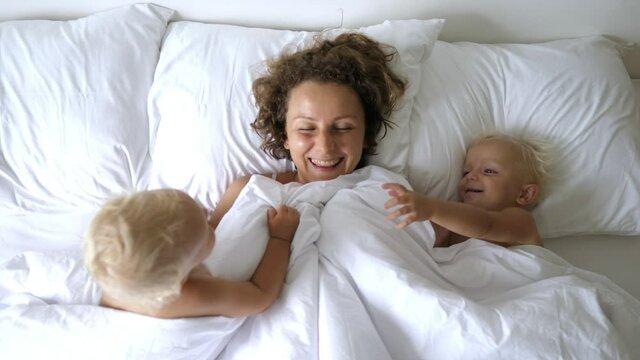 Top view of young caucasian mother playing in white bed with her twin babies. Long mornings in a loving family