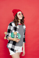 Happy stylish girl teenager wearing glasses, red beanie, white shorts and plaid shirt posing with penny board on red background